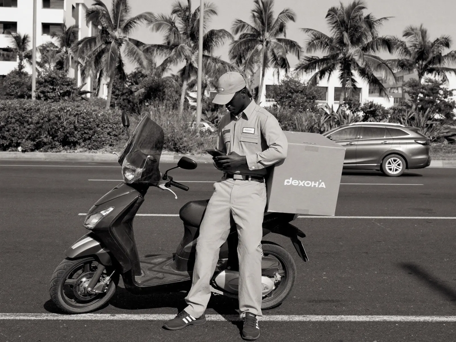 A courier reads his phone at the roadside, leaning against a scooter with a delivery box strapped to it.