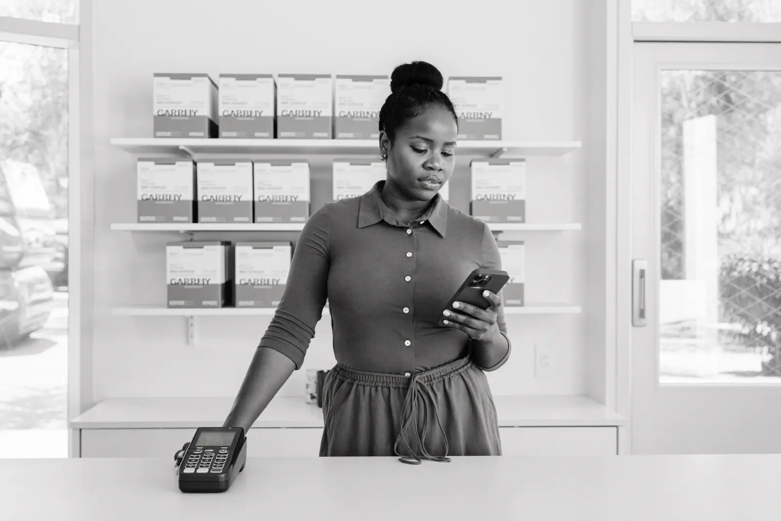 A shop owner checks her phone behind the counter of her store, a card payment terminal beside her.